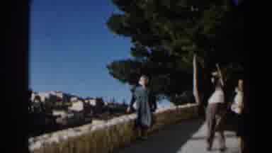 Israel (1960): Woman Wearing Grey Coat Walks Past White Stone Wall On A Sunny Day