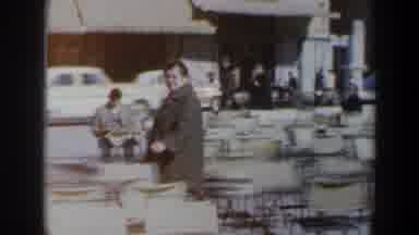 Israel (1960): Person Wearing Grey Coat Walks By White Chairs On A Sunny Day