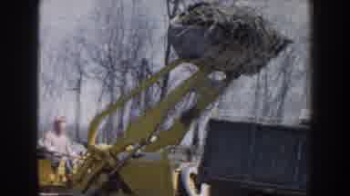 Nebraska (1959): Yellow Bulldozer Pours Dirt Into Blue Dump Truck On A Clear Day