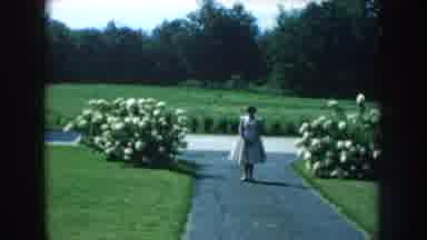 Nebraska (1959): Woman In White Dress Standing On Driveway Between Bushes With