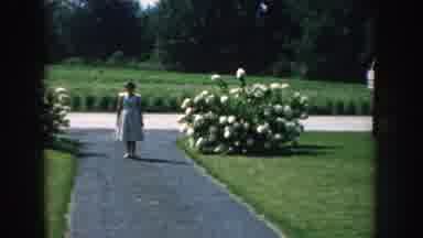 Nebraska (1959): A Woman In A White Dress Standing On A Small Path In A Garden