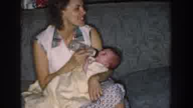 Nebraska (1959): Smiling Woman Feeds A Baby Wrapped In A White Blanket On A Sofa