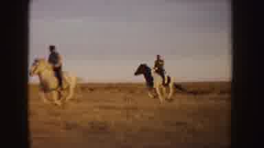 Idaho (1955): Two People Ride Horses Through Tan Field Towards Brown Hut On A