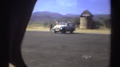 Sudan (1969): View Of White Car Parked On Brown Field And People Dancing On Beach