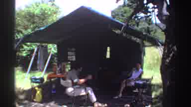 Nigeria (1969): Two People Make A Toast While Sitting Under Green Tent On A Sunny