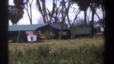 Kenya (1969): Panoramic View Of People Sitting In Tents Under Trees And Cars