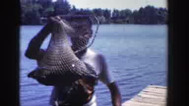 Wisconsin (1953): Man Wearing White Shirt Holds Grey Fish In Net On A Sunny Day
