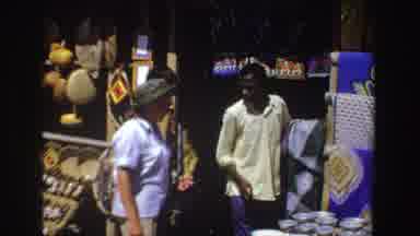 South Africa (1969): Woman Examines Crafted Items Shown By Man In Outdoor Bazaar