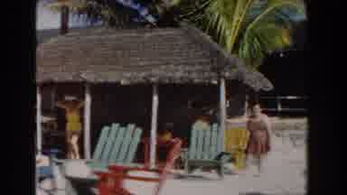 Nassau Bahamas (1961): Woman Wearing Yellow Pants Walks Out Of Tan Hut On A Bright