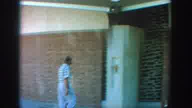 Wisconsin (1964): Man Wearing Blue Shirt Walks Into Tan Building On A Clear Day