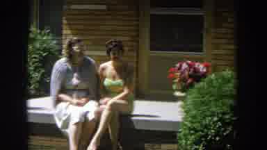 Wisconsin (1964): Two Women Sitting Near Tan Brick House On A Sunny Day