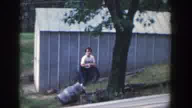 Wisconsin (1964): Girl Wearing Blue Pants Walks Gray Shed On A Clear Day