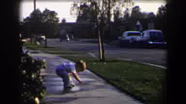 California (1958): Small Boy Wearing Gray Pants Touches Small Puddle On Sidewalk