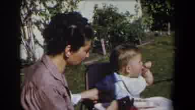 California (1958): Woman Wearing Brown Shirt Feeds Baby Wearing White Bib On A