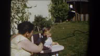 California (1958): Toddler Wearing White Shirt Is Fed By Woman Wearing Green Dress