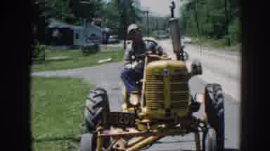 Nebraska (1958): Man Wearing Blue Pants Drives Yellow Tractor Down Street On A