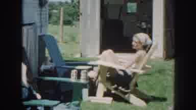 Nebraska (1958): Woman Wearing Gray Head Scarf Sits In Pale Yellow Wooden Chair On
