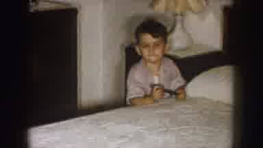 Nebraska (1957): Small Boy Wearing Tan Shirt Kneels Besides White Bed Holding