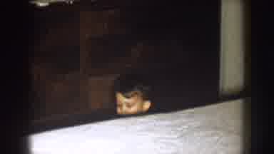 Nebraska (1957): Boy Wearing Tan Shirt Sits Behind White Bed Near Red Hanging