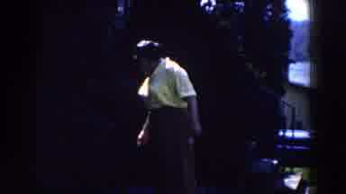 Michigan (1959): Woman Wearing Yellow Shirt Stands Near Dark Green Hedges On A