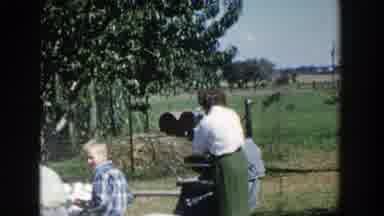Wisconsin (1958): Woman Wearing Green Skirt Stands Near People Eating At Picnic