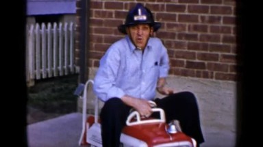 1965: Man Sitting on Toy Car with Helmet in Outdoor Setting