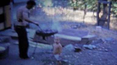 1973: Man grilling food outside trailer house for wife and baby.