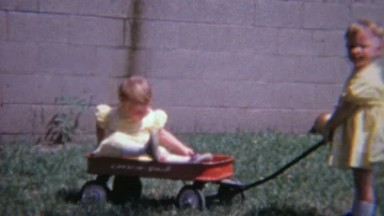 1965: Twin Toddler Girls Dressed Identically Riding Red Wagons Outdoors