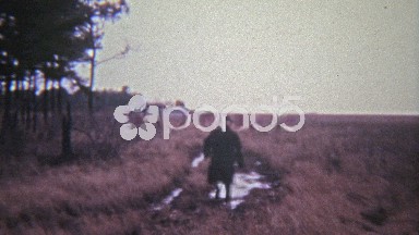 1972: Old man walking through mud puddle in open grass field.
