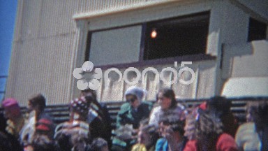 1963: Crowd seated watching high school track meet during bright summer day.