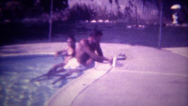 1972: Leathery suntan man smoking in pool with young bikini clad companion. SAN