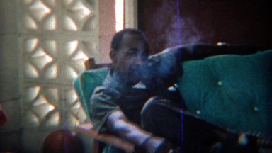 1969: Confident African man smoking cigarette outdoor couch party.
