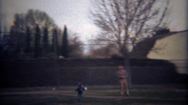 1979: Father son fly kite together in residential neighborhood park.