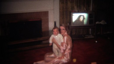 1979: Mother holding baby in front of background tv set.