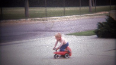 1979: Blonde young boy pedals play toy scooter around front yard.