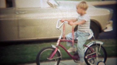 1979: Boy learning tricycle pedaling on suburban sidewalk.