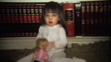 1974: Little Girl Playing with Doll in Front of Encyclopedia Book Set.