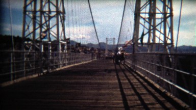 1972: People walking across bridge on windy day, flags blowing.