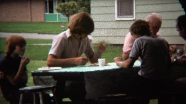 1973: Thick full 70's hair man eating at outdoor picnic event.