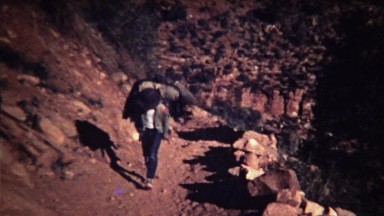 1972: Boy walking up dusty rocky mountain trail with backpack.