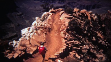 1972: Man hiking dry desert mountain trail with red backpack to river bed.