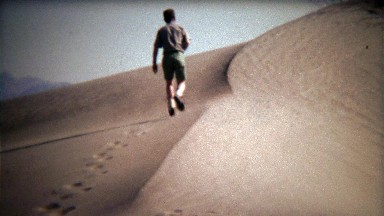 1972: Man walking up classic dry desert sand dunes.