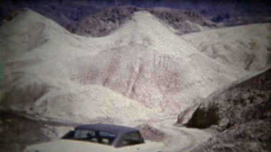 1972: Classic big car driving down steep hill in white desert dry wasteland.