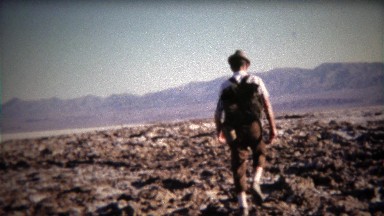 1971: Man walking across mountainous dry lake bed with backpack.
