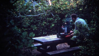 1971: Man lighting camp stove on picnic table in forest wilderness.