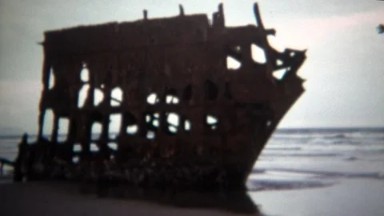 1971: Rusty broken old shipwreck beached on sandy ocean shore.
