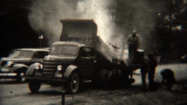 1939: Asphalt road laying truck construction workers shoveling.