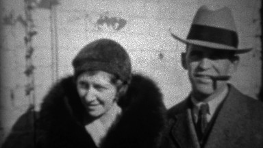 1933: Fedora hat man smoking cigar with loyal wife in black fur coat.