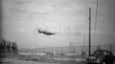 1938: Goodyear lifeguard tire blimp flying above classic car.