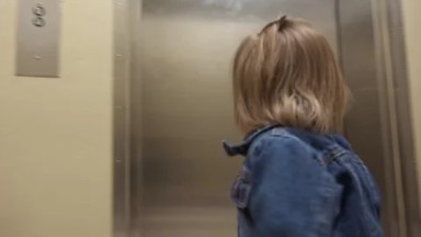 Mom holding elevator door for toddler entering hospital hallway.